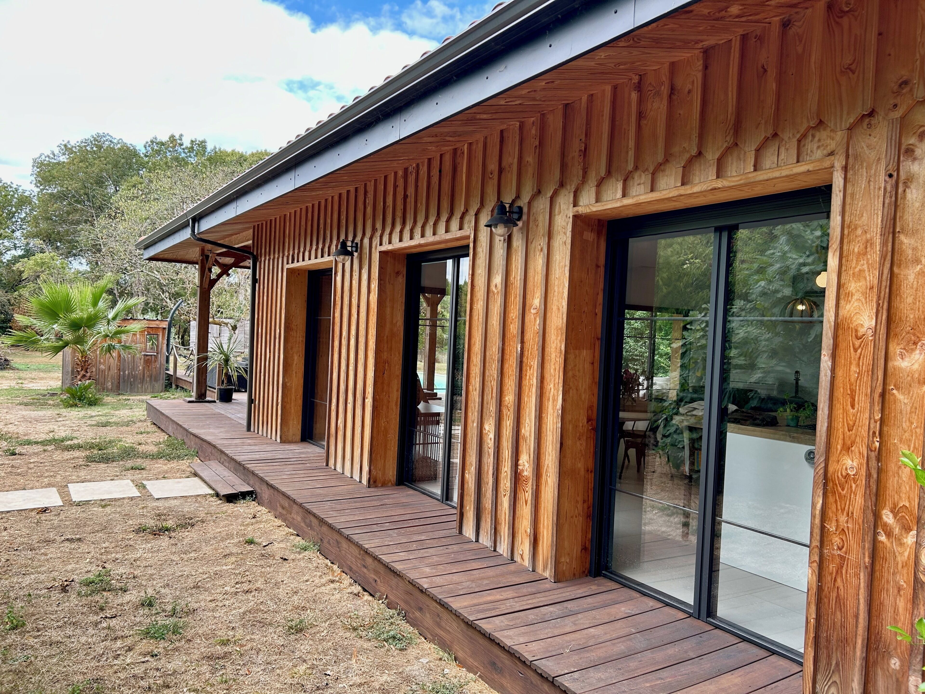 Une façade en bois, de grandes portes-fenêtres coulissantes, une terrasse en bois et un petit espace extérieur agrémenté d’un palmier.
