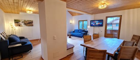 Un amplio salón con techo de madera, un sofá azul, una mesa de comedor con sillas y un televisor montado en la pared.