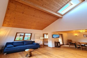 Een ruime woonkamer met een hoge plafond, houten vloeren, een blauwe bank, een klein rond tafeltje en een aan de muur bevestigde televisie.