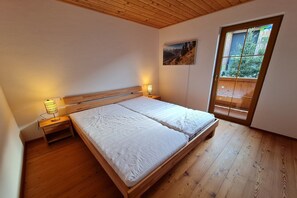 A bedroom with a wooden bed, bedside lamps, a window with a view of greenery, and a painting on the wall. - Modern apartment in Bad Kleinkirchheim (Bad Kleinkirchheim)