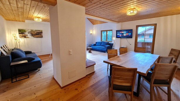 Ett rymligt vardagsrum med ett träpanelat tak, en blå soffa, ett matbord med stolar och en platt-TV som är monterad på väggen.