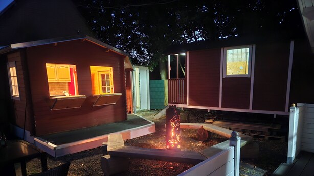 A wooden cabin with a fire pit in the center, surrounded by a deck and a small garden area.