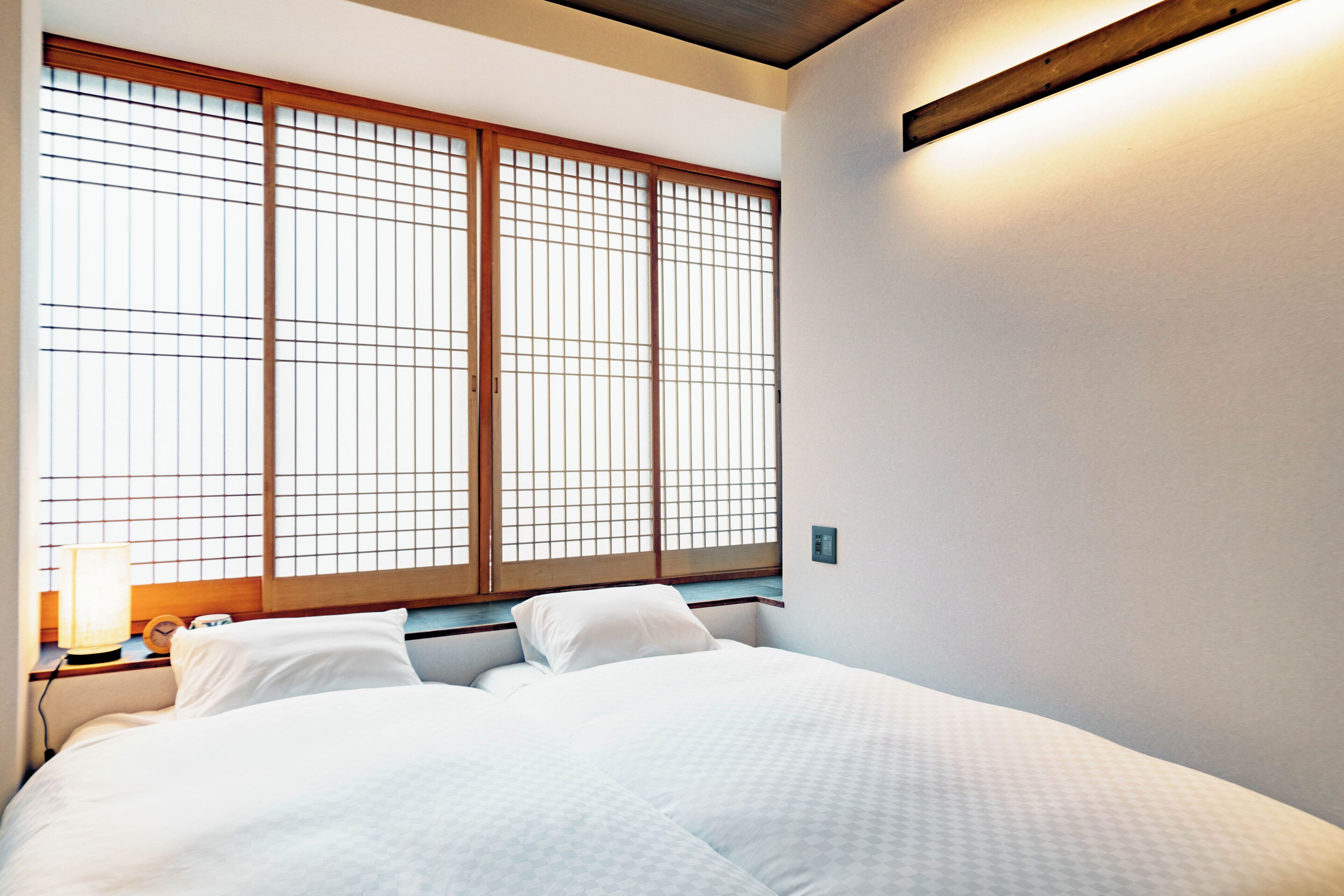 Habitación de hotel con cama, mesita de noche, lámpara y un ventanal grande con marcos de madera.