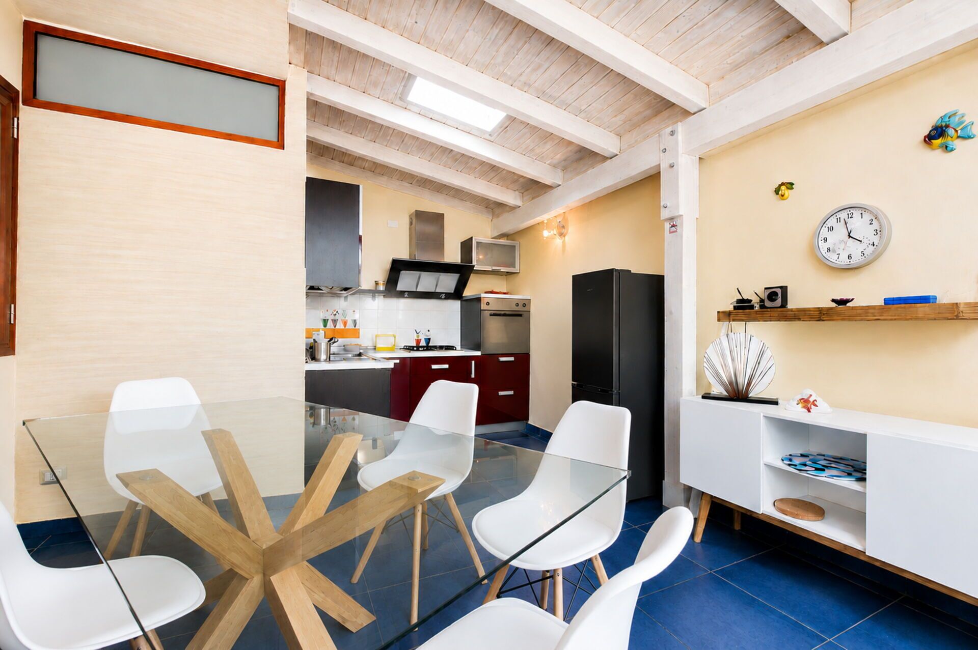Une cuisine moderne avec un coin repas, comprenant une table en verre et des chaises blanches, un réfrigérateur noir et une horloge murale.