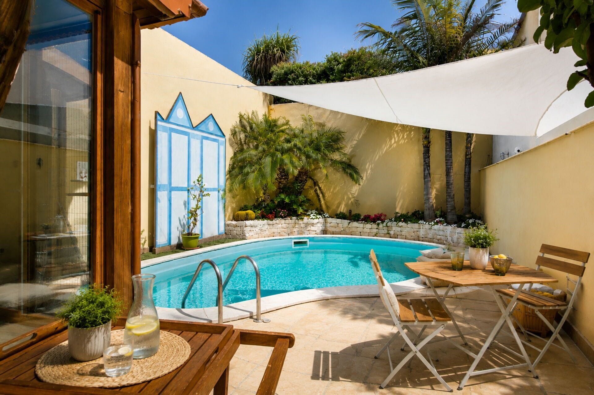 A pool area with a wooden table and chairs, a white canopy, and a stone wall with plants.
