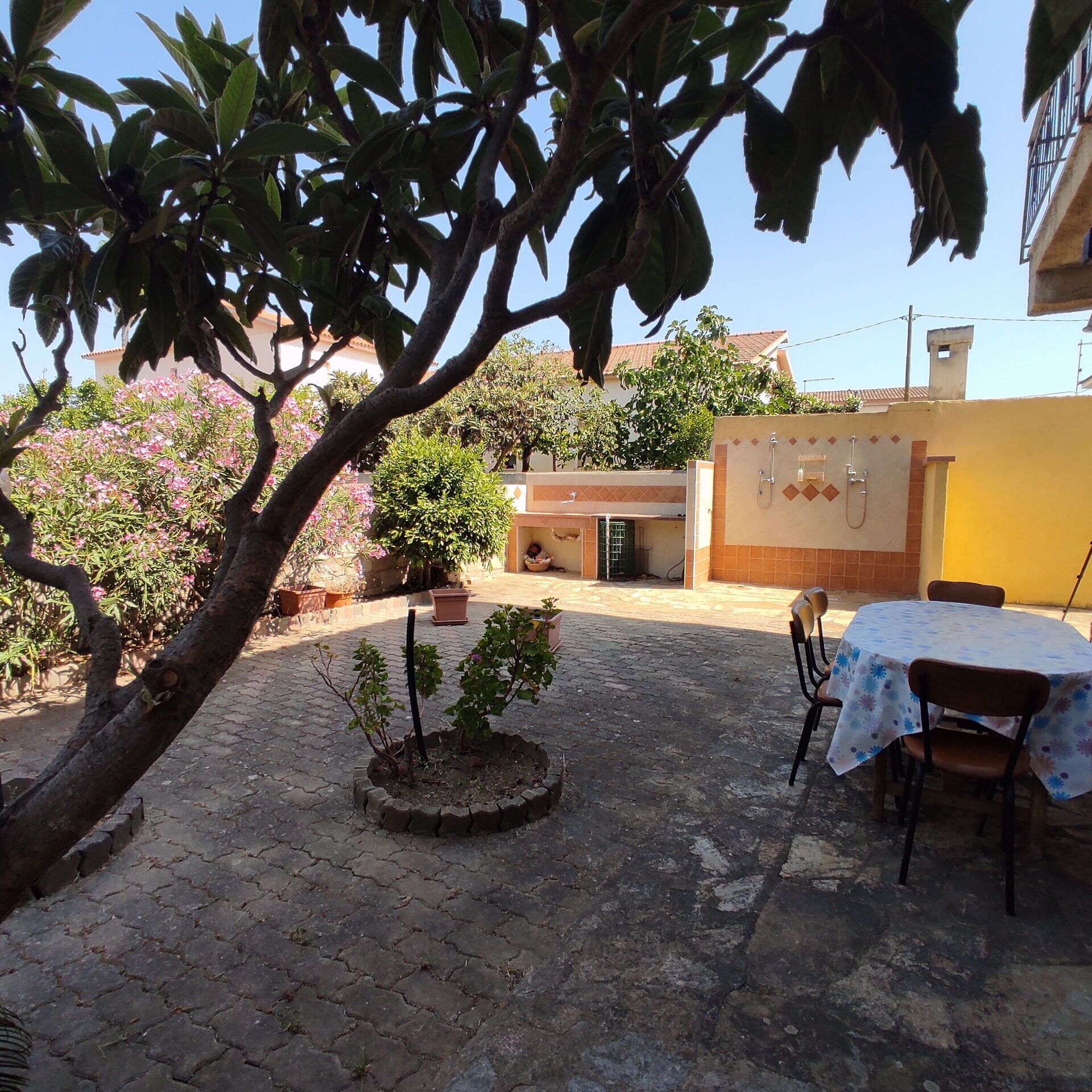 Une cour avec un sol carrelé, un petit arbre, une plante en pot, une table avec des chaises et un bâtiment dont un mur est recouvert de carreaux et qui possède une porte.
