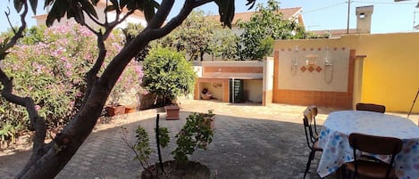 A courtyard with a tiled floor, a small tree, a potted plant, a table with chairs, and a building with a tiled wall and a door.