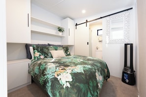 Un dormitorio con cama, una estantería con una planta, un armario y una aspiradora.