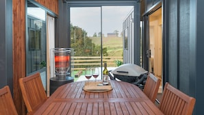 Eine überdachte Terrasse mit Tisch für zwei Personen, einem Grill und einer Weinflasche.