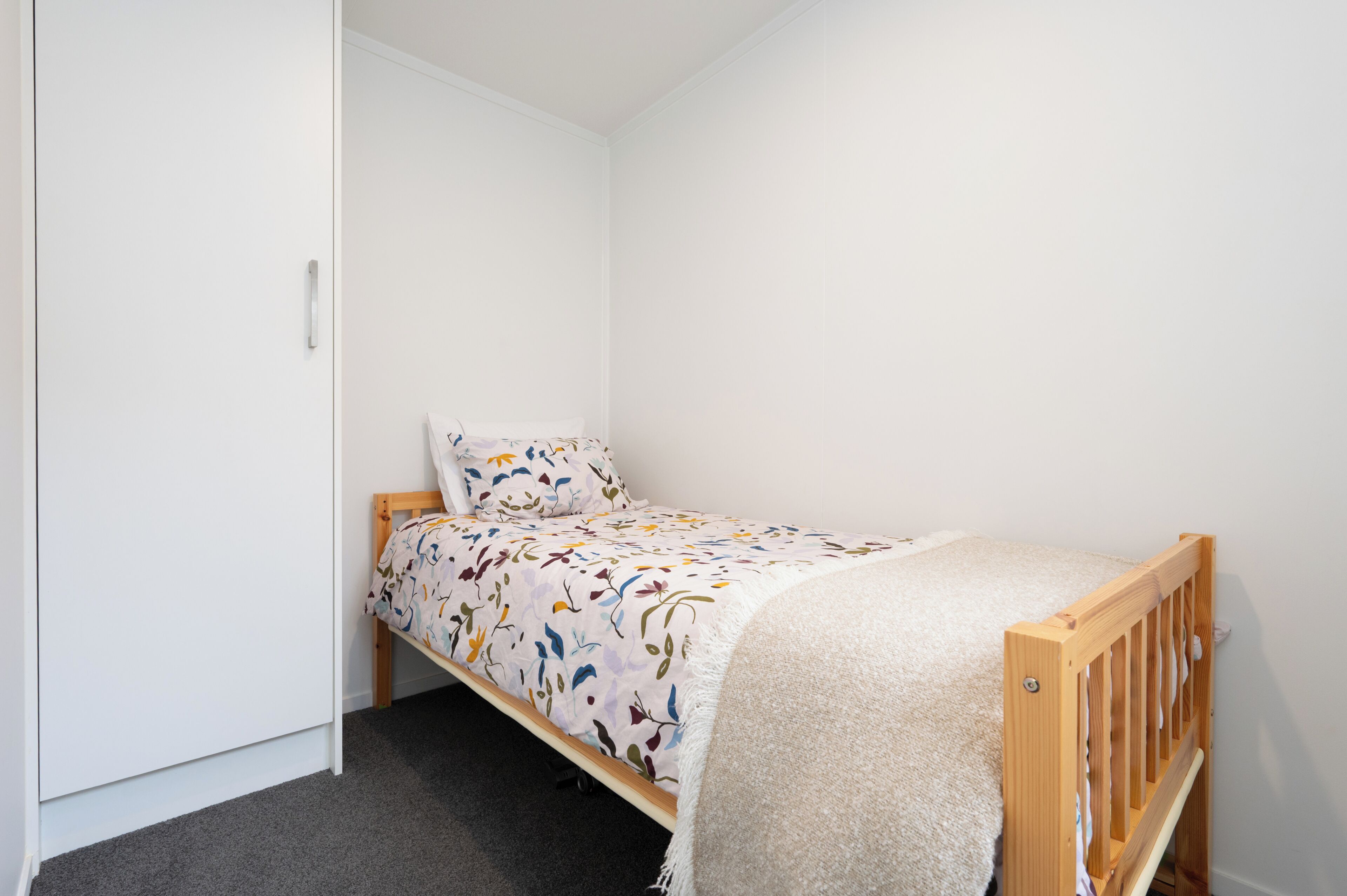 A small, single-bed room with a wooden frame, a white wall, and a grey carpet.