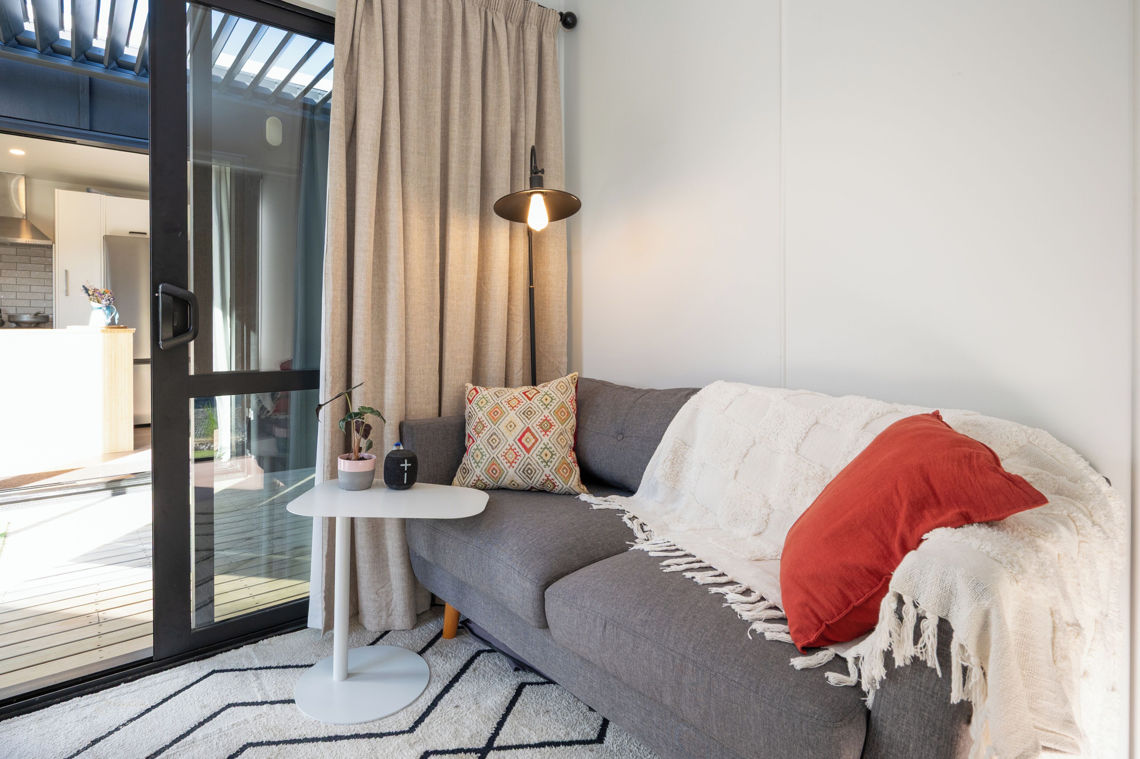 A modern living room with a grey sofa, a small white side table, a potted plant, and a red pillow.