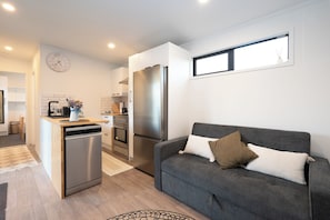 A modern kitchen with a dishwasher, oven, and refrigerator. There is a grey sofa with cushions and a window above it. - Haven Hills Retreat - Romantic, luxury off grid tiny house with stunning views. (Taumarunui)