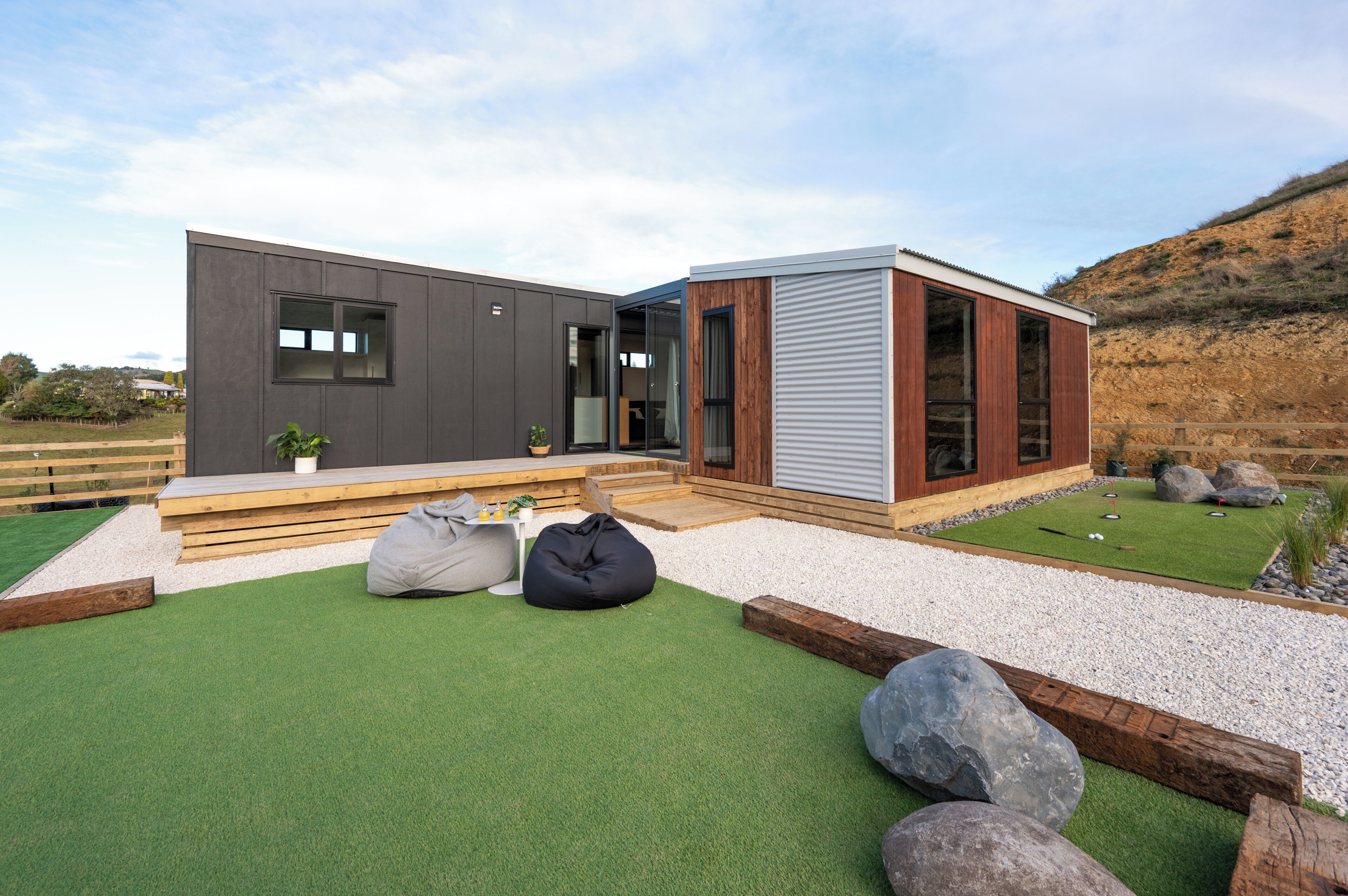 Une maison moderne à l’extérieur gris, dotée d’une terrasse en bois et d’un petit jardin agrémenté de gazon synthétique et de rochers.
