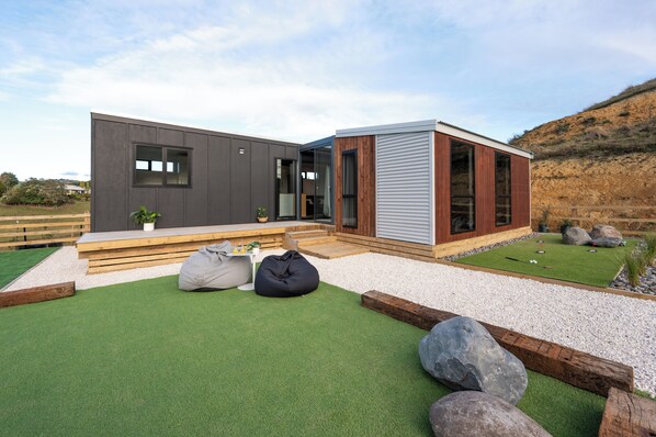 Una casa moderna con fachada gris, terraza de madera y un pequeño jardín con césped artificial y rocas.