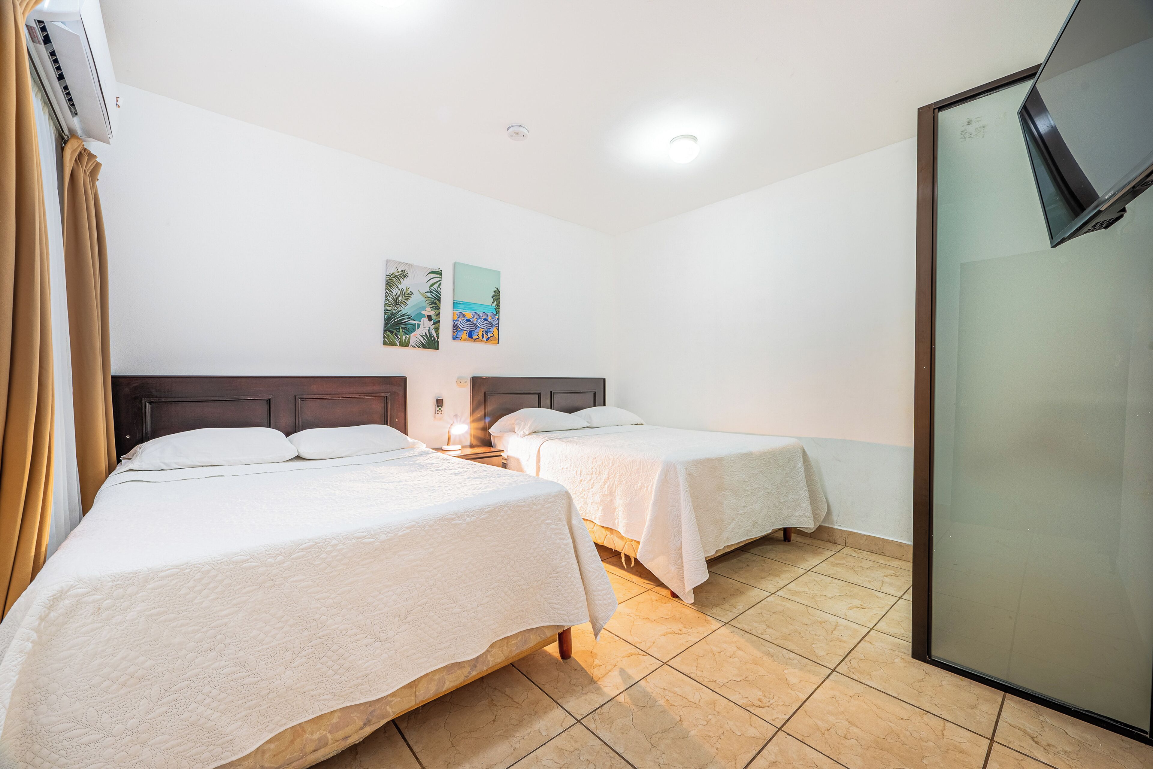 Quarto de hotel com duas camas, uma janela com cortinas, ar condicionado fixado na parede e duas obras de arte na parede.