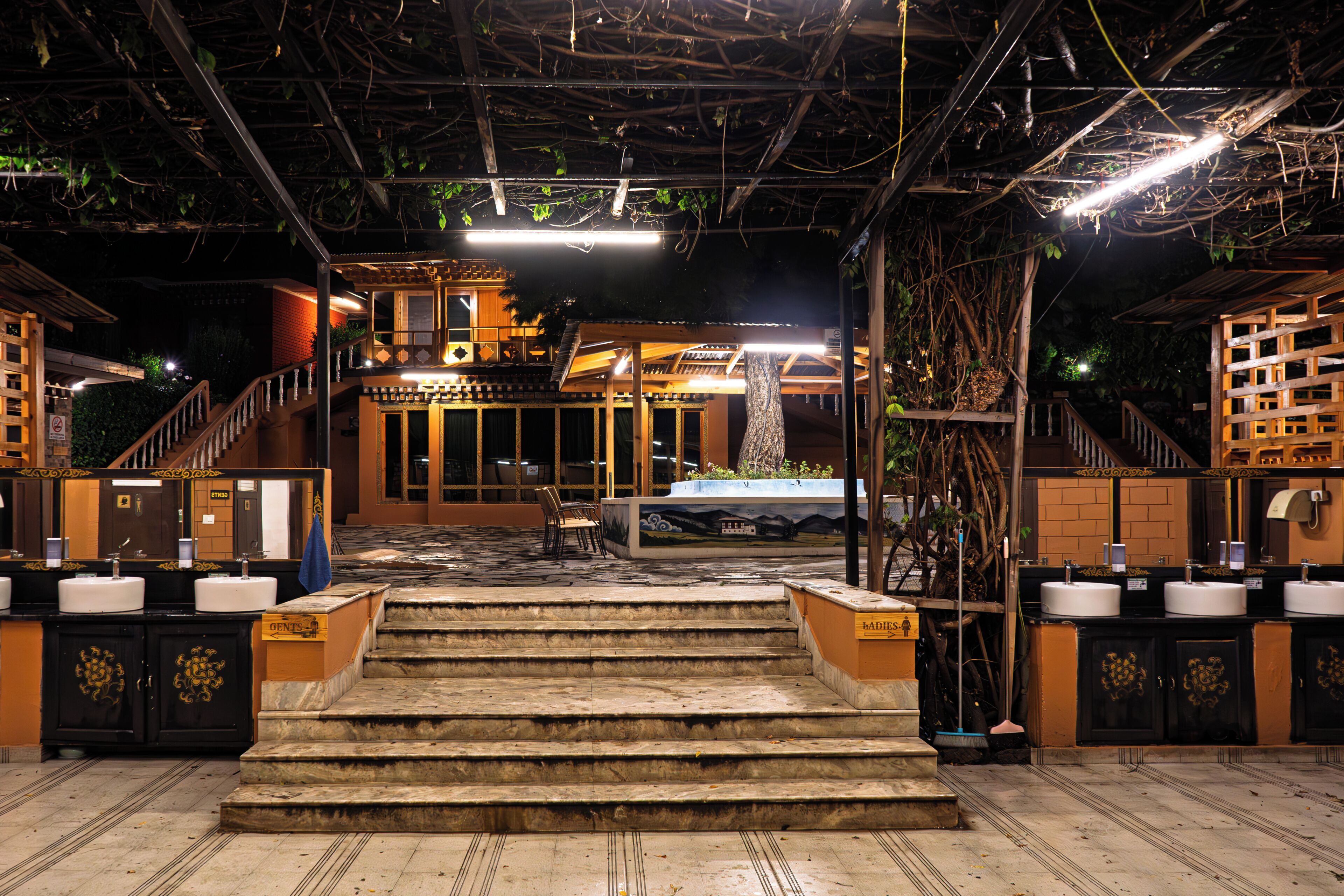 A hotel lobby with a staircase leading to a second-floor area, a reception desk, and a large tree in the center.