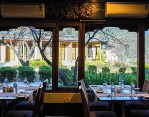A dining area with tables set for two, large windows with a view of a building and trees, and air conditioning units on the ceiling.