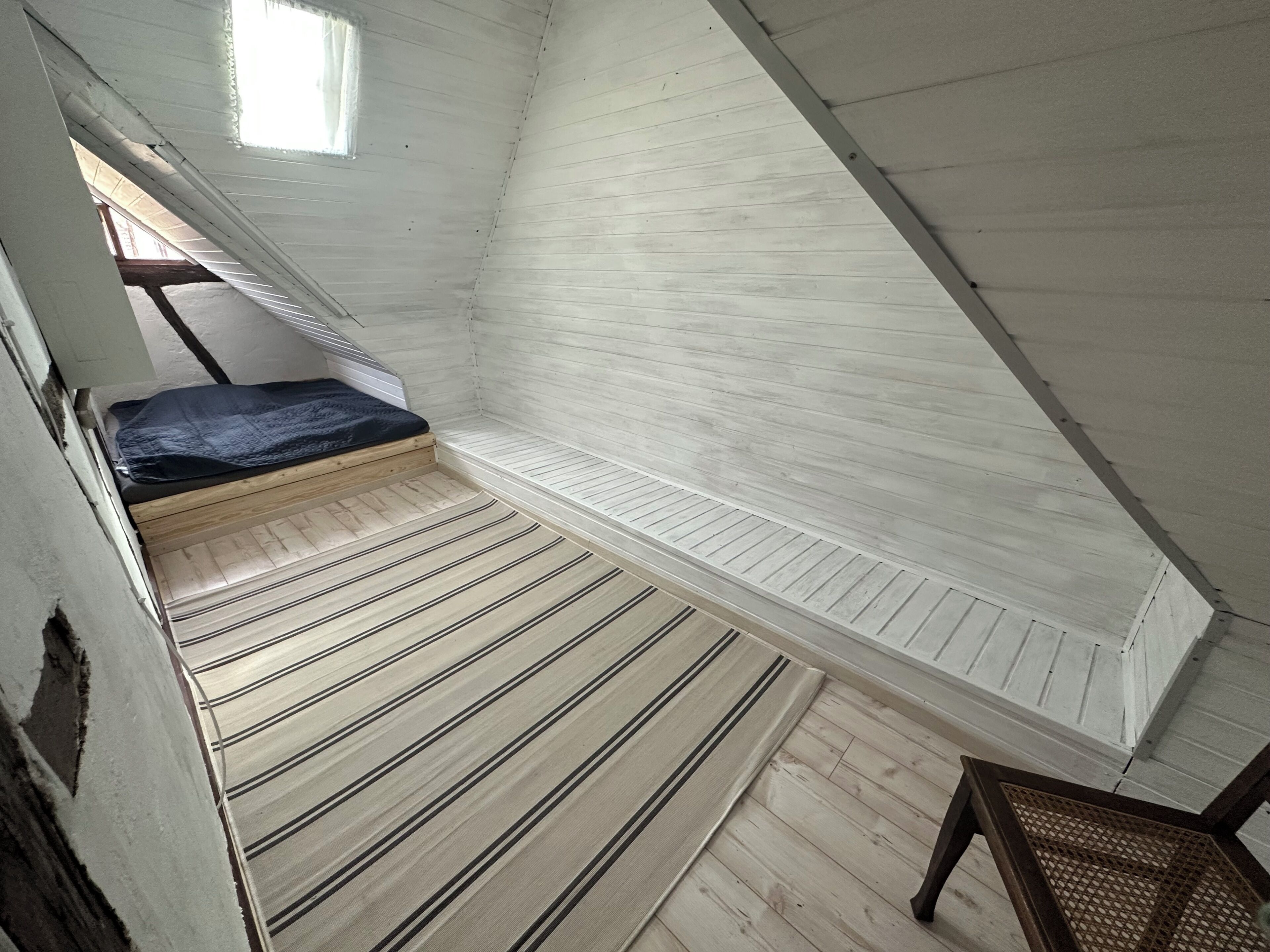 Ein Zimmer mit schrägverlaufender Decke, einem Einzelbett mit dunkler Bettdecke und einem Holzboden mit Streifenmuster.