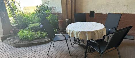 An outdoor patio with a round table and chairs, surrounded by potted plants and a brick floor.
