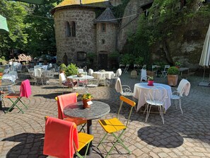 An outdoor café with stone walls, tables, and chairs, surrounded by greenery.