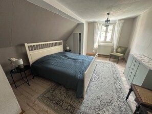 A bedroom with a bed, a nightstand, a chair, a dresser, and a window with curtains.