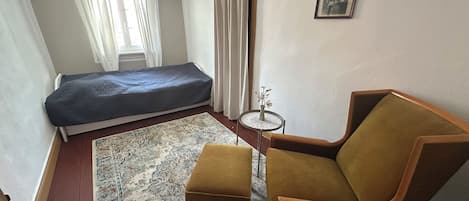 Une petite pièce meublée, comprenant un lit, une chaise, une petite table et un tableau accroché au mur.