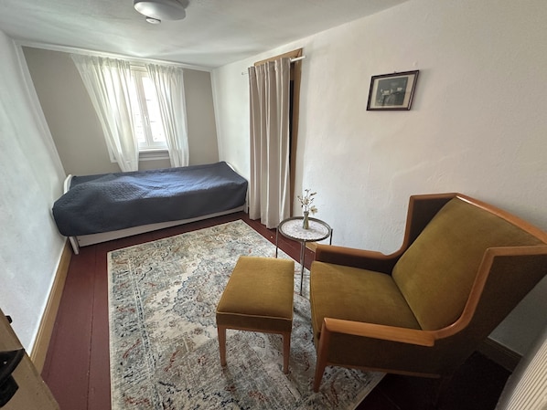Una stanza piccola e arredata con un letto, una sedia, un tavolino e un quadro appeso al muro.