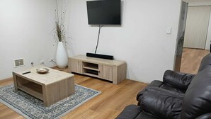 A living room with a wooden coffee table, a black leather sofa, a TV on the wall, and a white vase with plants.