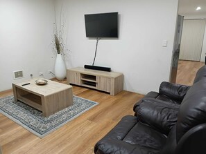 A living room with a wooden coffee table, a black leather sofa, a TV on the wall, and a white vase with plants.
