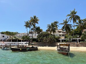 A beachfront with two boats docked, palm trees, and buildings in the background.