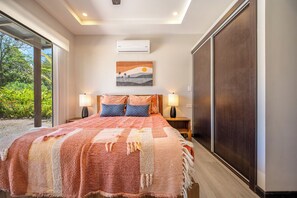 Un dormitorio con una cama grande, dos lámparas de noche, un aire acondicionado de pared y una ventana con vistas a zonas verdes.