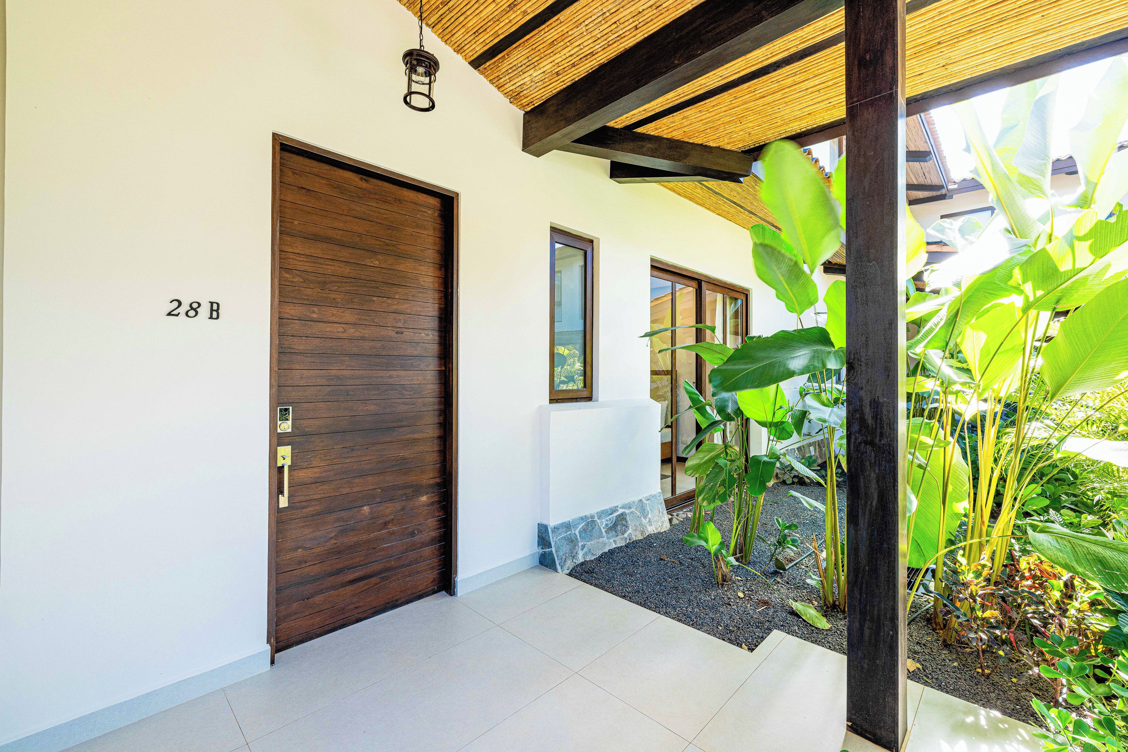 A modern entrance with a wooden door marked '28B', a tiled floor, and a small garden area with tropical plants.