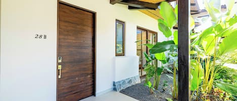 A modern entrance with a wooden door marked '28B', a tiled floor, and a small garden area with tropical plants.