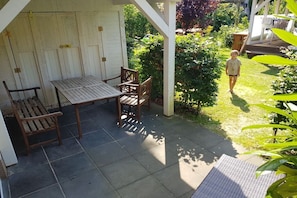 A covered patio area with a table and chairs, surrounded by a garden with a child walking. - Holiday apartment with 2 bedrooms (Kölpinsee)