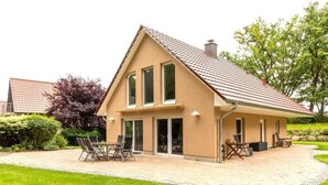 Ein zweistöckiges Haus mit geneigtem Dach, einer Terrasse mit Gartenmöbeln und einem gepflegten Rasen.