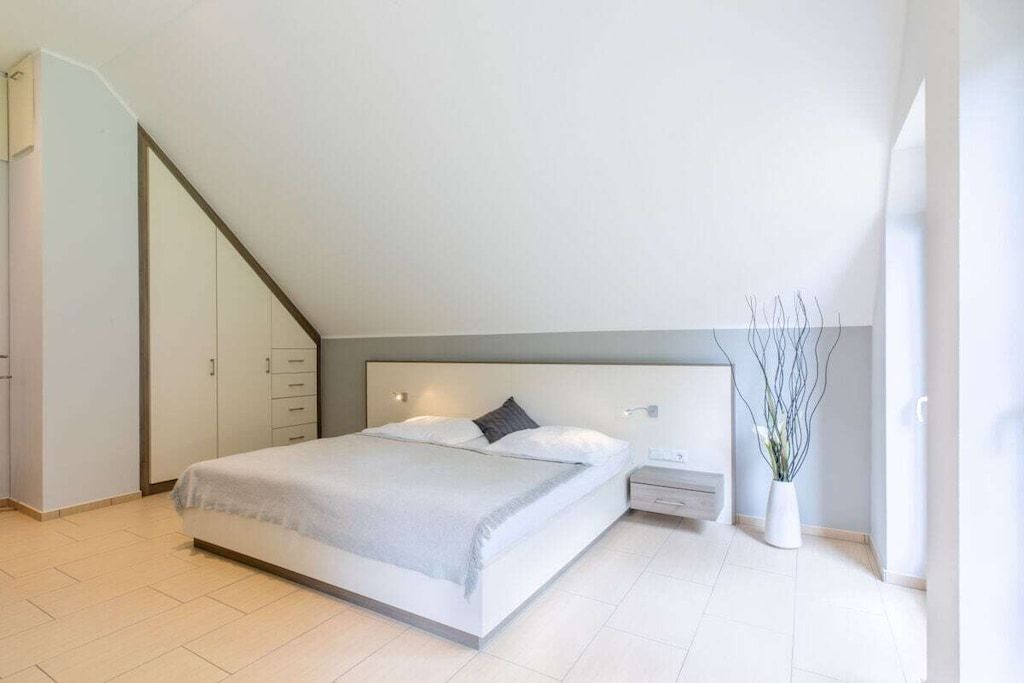 A modern bedroom with a bed, bedside tables, a nightstand, a wall-mounted lamp, and a potted plant.