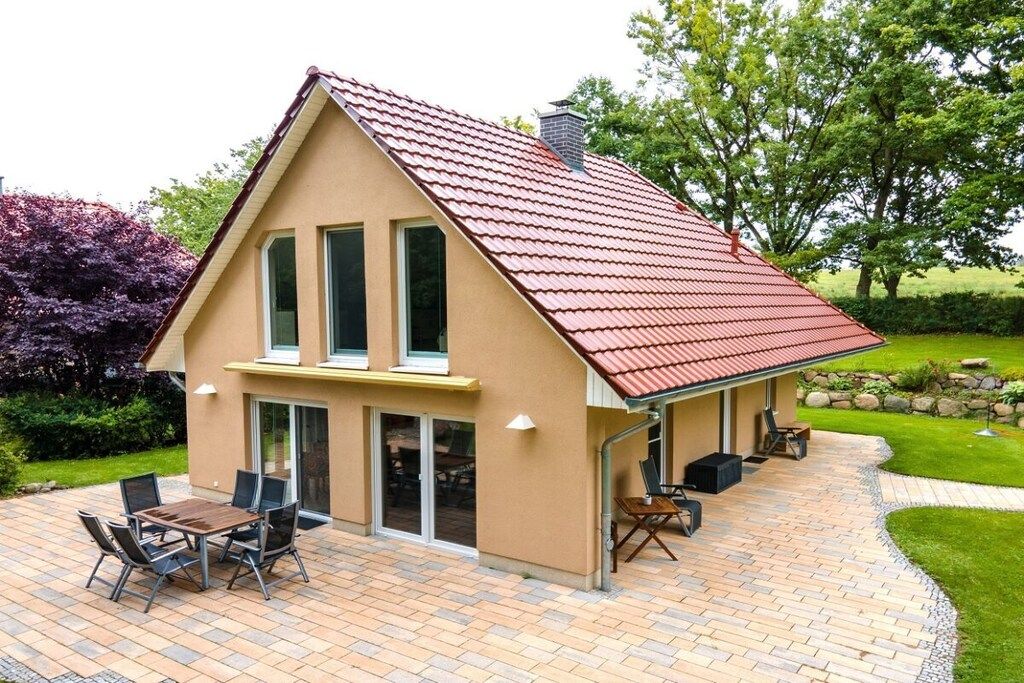 A house with a red roof, a patio with a table and chairs, and a garden with trees and shrubs.
