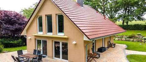A house with a red roof, a patio with a table and chairs, and a garden with trees and shrubs.