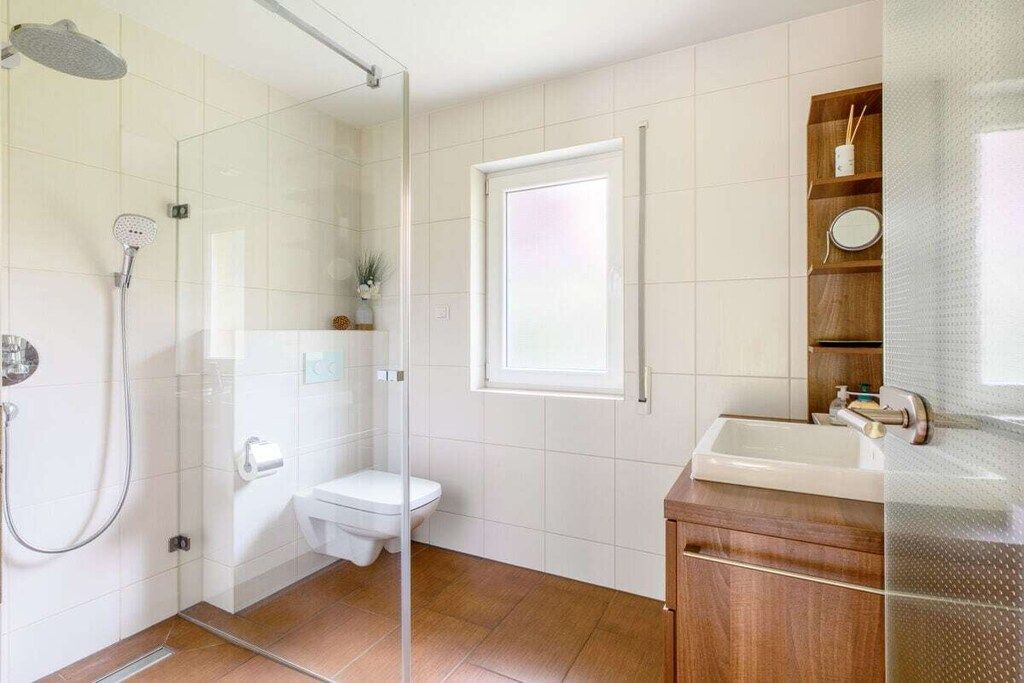 Ein modernes Badezimmer mit Duschkabine aus Glas, Toilette, Waschbecken mit Holzschrank und einem Fenster für natürliches Licht.