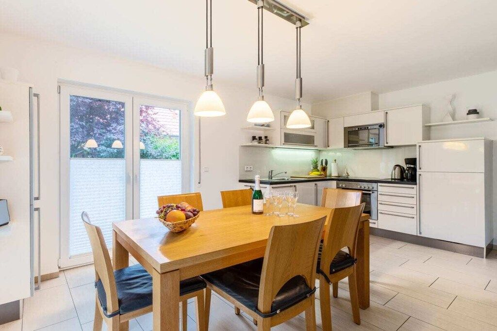 Una cocina moderna con armarios blancos, una mesa de comedor con sillas e iluminación colgante.