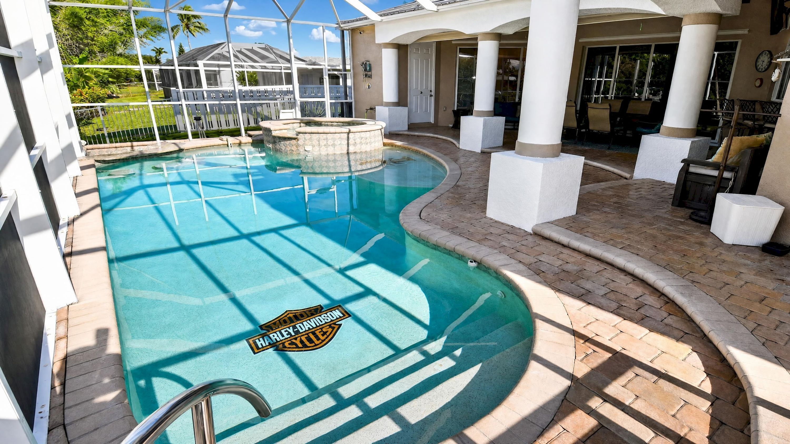 A swimming pool with a Harley-Davidson logo, surrounded by a covered patio area with white columns and outdoor furniture.