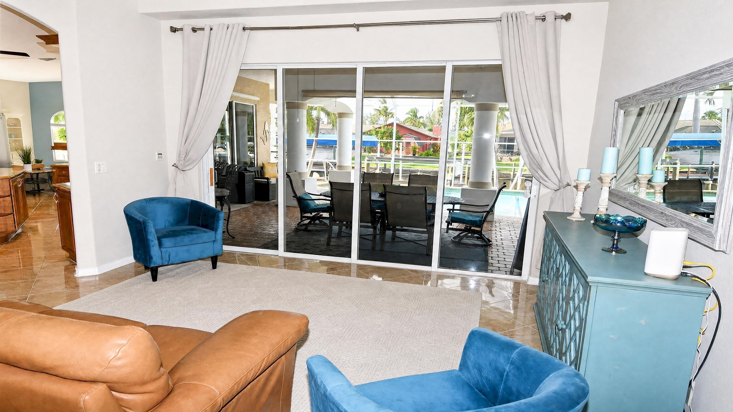 Un salón con un sofá de cuero marrón, un sillón azul y un sofá azul. Hay una puerta de vidrio que da a un patio exterior con una mesa y sillas.