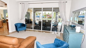 A living room with a brown leather sofa, a blue armchair, and a blue sofa. There is a glass door leading to an outdoor patio with a table and chairs. - Almare Gulf Access Pool Spa Boat Dock (Cape Coral)