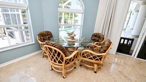 A dining area with a glass-top table surrounded by wicker chairs, a view of a pool through a window, and a tiled floor. - Almare Gulf Access Pool Spa Boat Dock (Cape Coral)