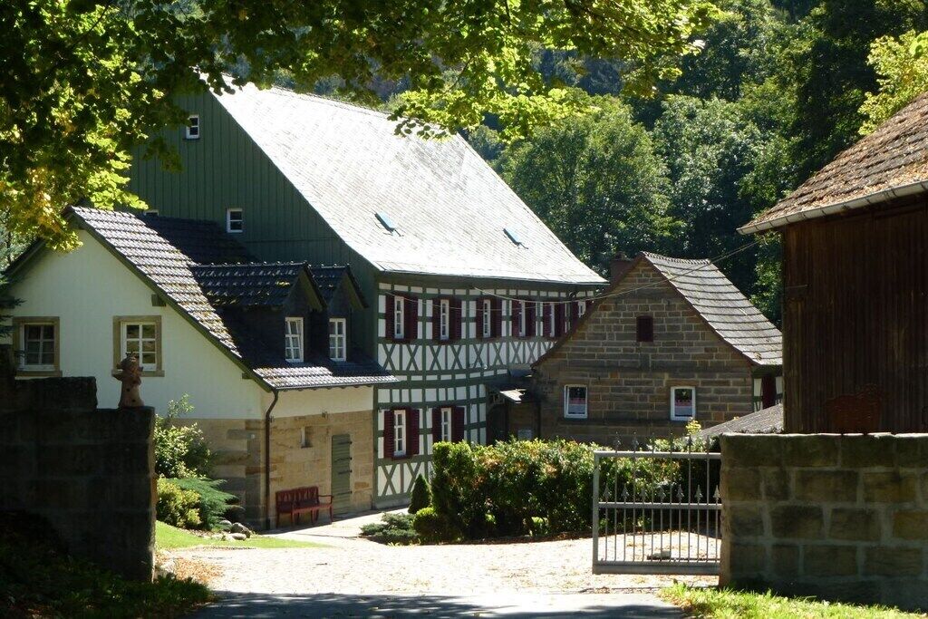 Ein traditionelles Fachwerkhäuschen neben einem Gebäude aus Stein, eine Schotterauffahrt und ein Metalltor.