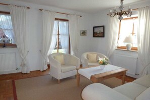 Ein Zimmer mit zwei weißen Sesseln, einem Holztischchen und einem Sofa. Weiße Vorhänge sind an den Fenstern, es gibt eine Kristallleuchter und ein Bild an der Wand.