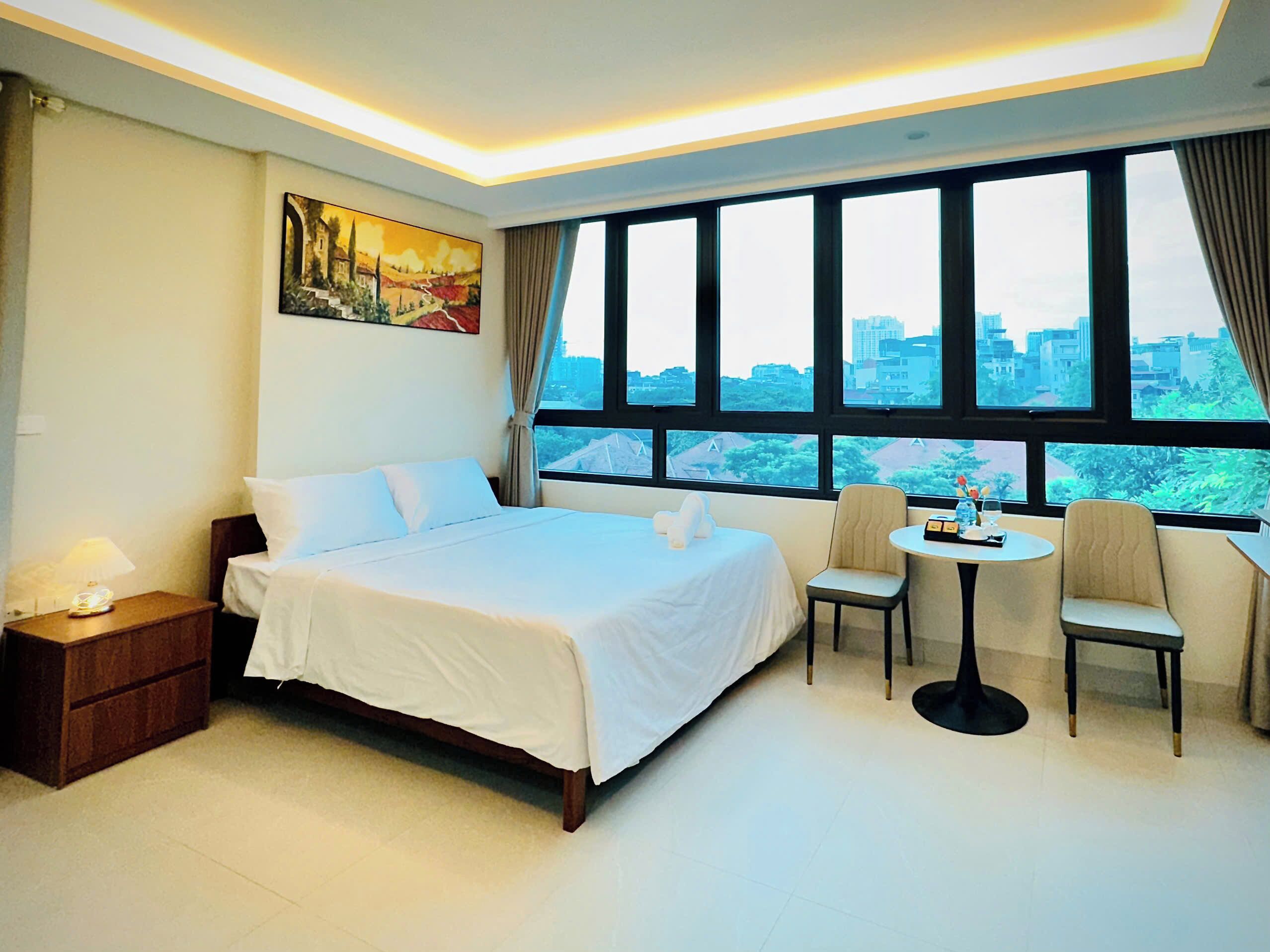 Une chambre d’hôtel avec un grand lit, une petite table avec des chaises, un tableau au mur et une vue sur la ville grâce à de grandes fenêtres.