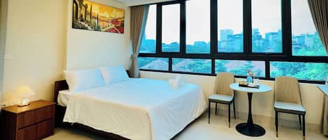 A hotel room with a large bed, a small table with chairs, a painting on the wall, and a view of the city through large windows.