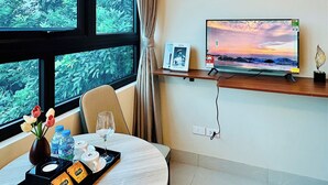 Habitación de hotel con un ventanal grande, un televisor de pantalla plana sobre un pedestal, una mesita con un surtido de artículos de aseo y un jarrón con flores.