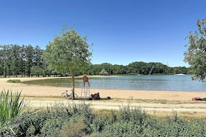 A lakeside setting with a bicycle, a ladder, and a person sitting on the sand.
